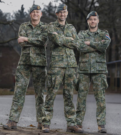 3 militairen in hetzelfde tenue poseren met de armen over elkaar met op de achtergrond een grote struik.