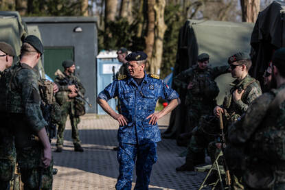 Een militair in blauw tenue staat met handen in zij tussen militairen in groen tenue.