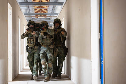 Vier militairen lopen met wapen gericht schouder aan schouder door een gang.