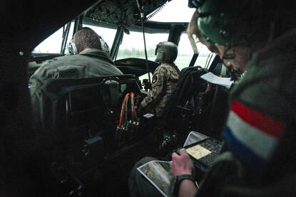 Drie personen in de simulator van een Chinook. Ze zitten in de cockpit. De persoon achter de twee vliegers heeft een scherm en kaart in zijn handen.