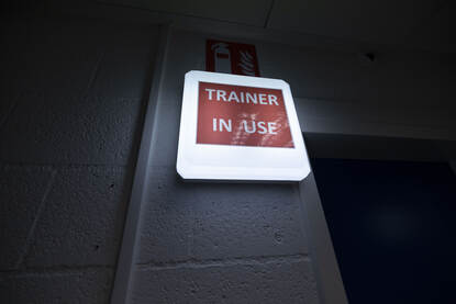 Vier afbeeldingen. Op een daarvan is een oplichtend bordje te zien met de tekst ‘trainer in use’. De andere drie foto’s tonen sergeant Michiel in de simulator en de virtuele wereld waarop hij uitkijkt vanuit de cabine van de Chinook. Ook is het boordwapen waarmee hij in de simulator werkt zichtbaar.