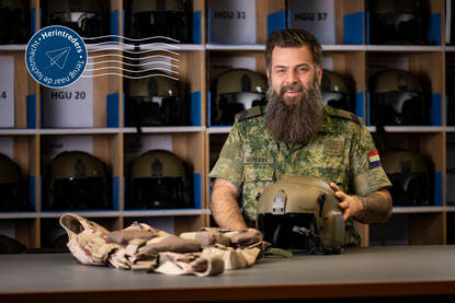 Sergeant Einar Nordsiek poseert achter een tafel met daarop een vliegerhelm en -vest. Achter hem een vakkenkast met vliegerhelmen.
