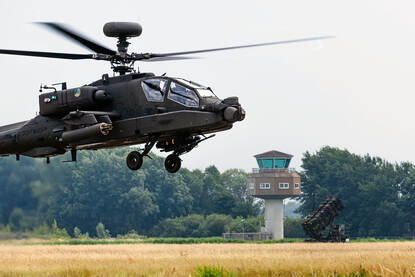 Gevechtshelikopter vliegt vlak boven de grond met op de achtergrond een verkeerstoren en Patriot-luchtverdedigingssyteem.