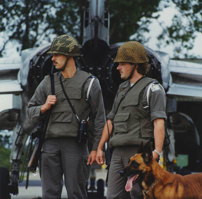 Twee militairen met een diensthond voor de achterkant van een straaljager.