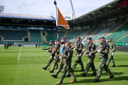 De vaandelwacht van de luchtmacht betreedt het veld vanuit een hoek van het stadion. De vaandeldrager in het midden, daaromheen militairen die een Colt C8-geweer vasthouden.