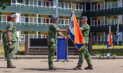 Een militair met een vlag in zijn handen staat tegenover een militair die de vlag in ontvangst gaat nemen.
