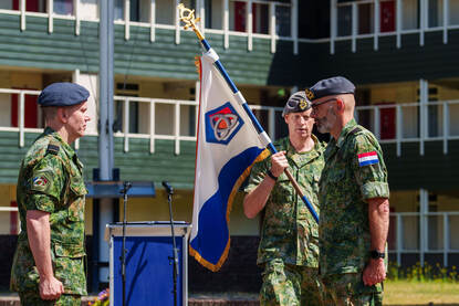 Twee militairen staan tegenover elkaar in de houding. Een derde militair heeft een vlag in zijn handen.