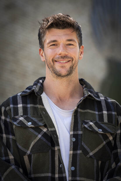 Man met stoppelbaard en geruit hemd kijkt lachend in de camera.