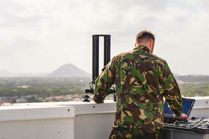 Militair met radiozendmast op dak met uitzicht op Aruba met in de verte een heuvel.