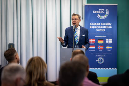 Sprekers geven een presentatie over de veiligheid van infrastructuur op de zeebodem voor een zaal met publiek.