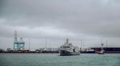 links: Twee grijze marineschepen varen op groen, golvend water. Het grootste schip heeft M940 op de zijkant, het kleinere M921, met antennes en apparatuur bovenop. Foto rechts: Een groot grijs marineschip vaart een haven binnen, met kranen, containers, en een bewolkte lucht op de achtergrond. Het water is kalm.