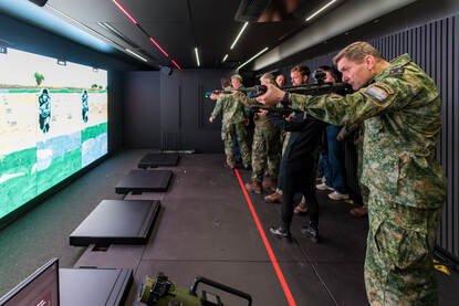 Militairen in uniform oefenen in een schietsimulator met virtuele projectie, ze richten met een wapen op een scherm met digitale doelen. Ze staan in rij.
